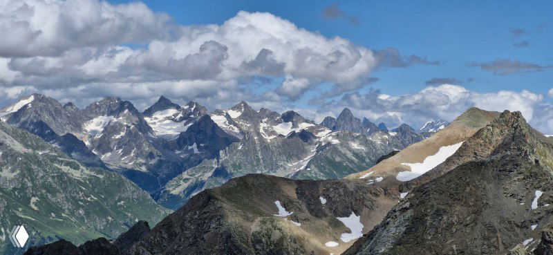 Широкий горный хребет с несколькими заметными вершинами и седловинами, на горизонте цепочка пологих склонов и облака.