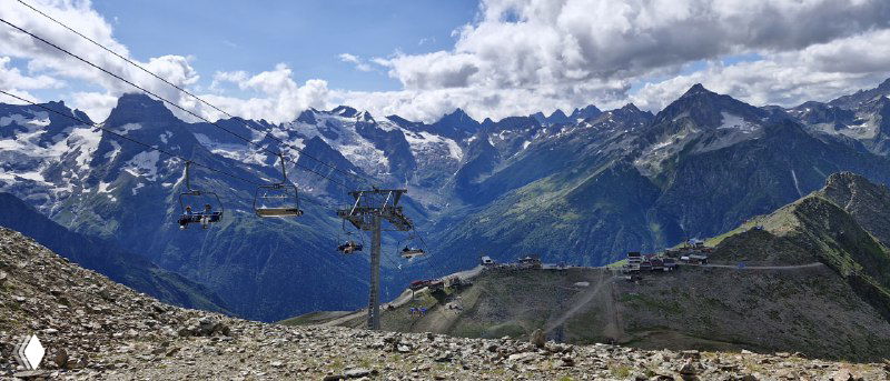 Канатные опоры и станция на переднем плане, далеко виднеются высокие горы и вершины Кавказа под переменной облачностью.