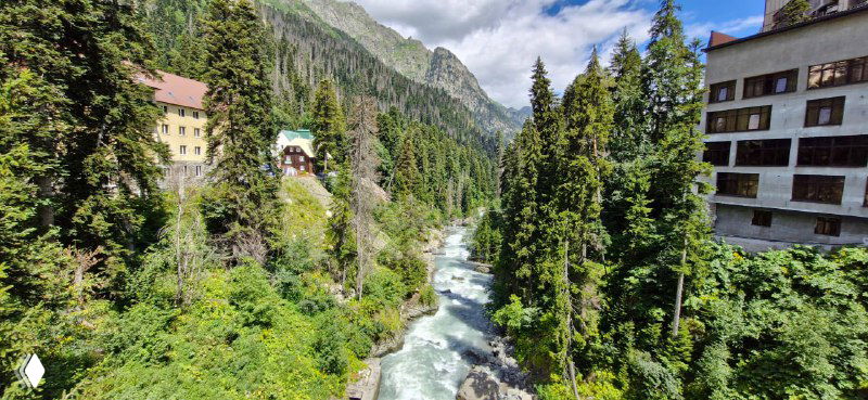 Горная река в узкой долине Домбая между склонами и хвойными деревьями; на правом берегу видны дома и гостиницы курорта.