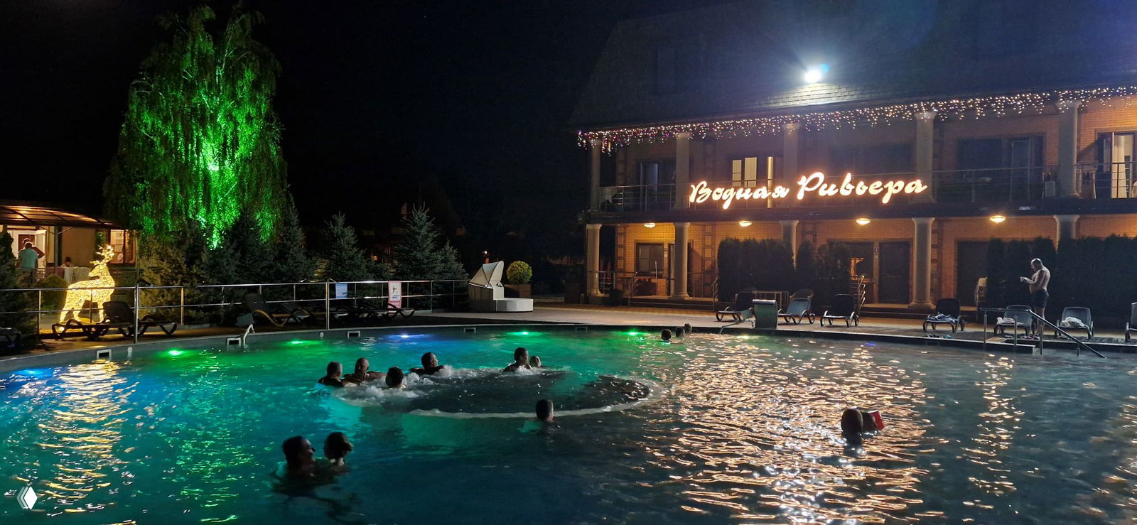 Ночной крупный бассейн с подсветкой и надписью «Водная Ривьера» на здании, люди купаются в тёплой воде при огнях.