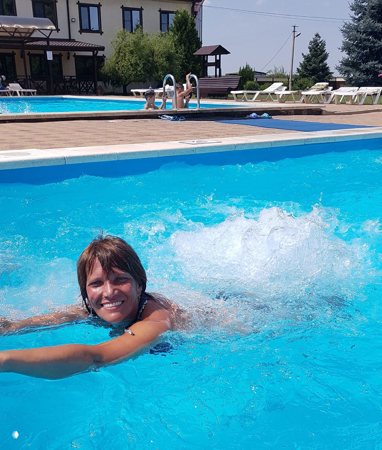 Дневной кадр: женщина улыбается в горячем бассейне, видны пузырьки гидромассажа и яркая голубая вода, летний солнечный день.