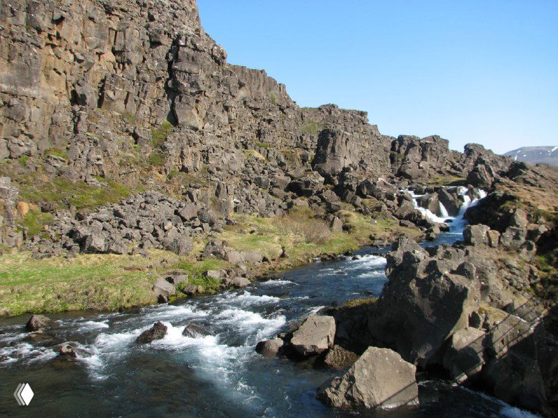 Горный поток, протекающий через лавовое поле и базальтовые скалы: пороги, пенящиеся струи и контраст чёрной лавы с бирюзовой водой.