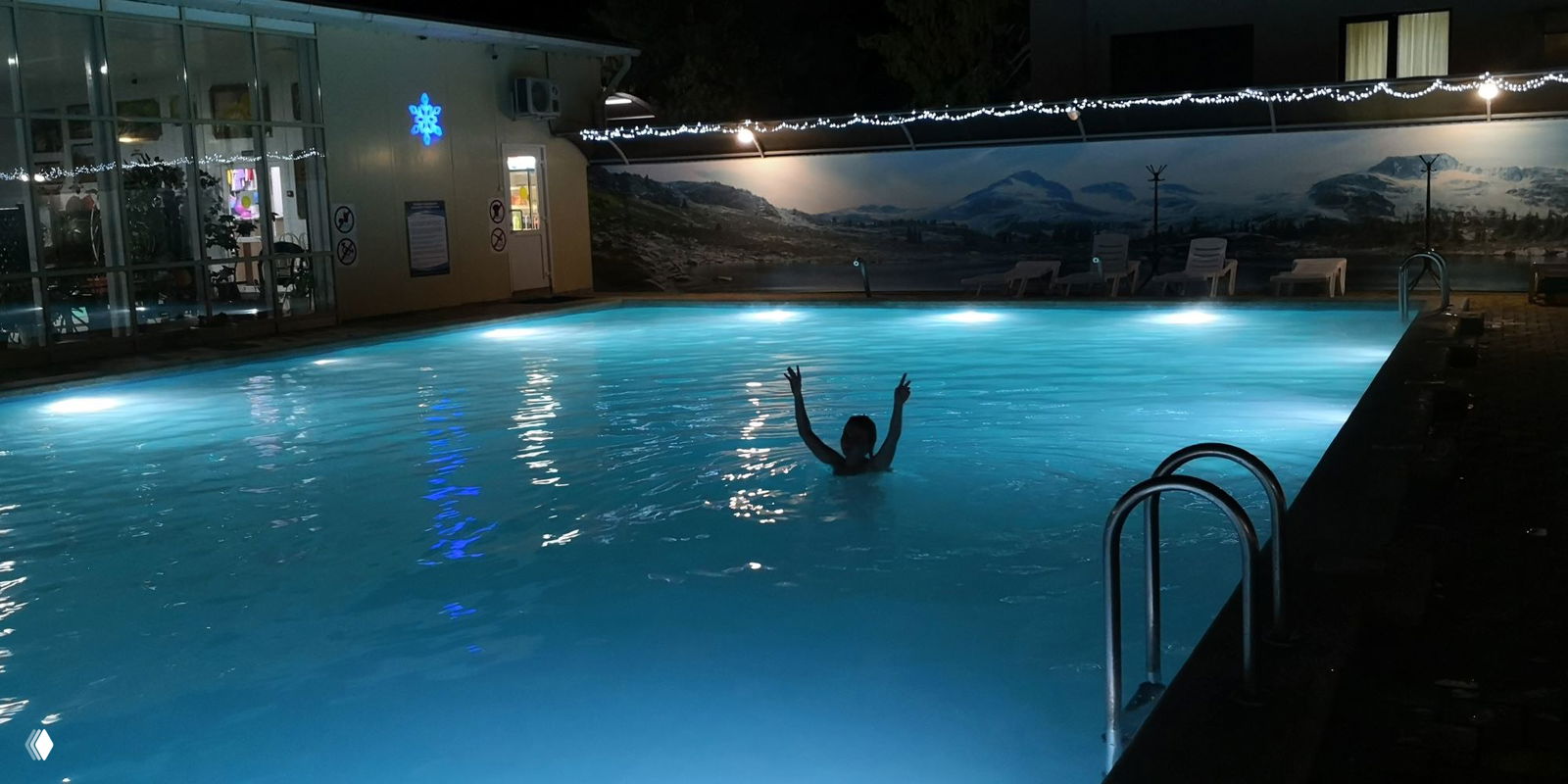 Ночной открытый бассейн с мягкой подсветкой, силуэт купающегося на фоне оформленной стены и тёплое освещение вокруг.