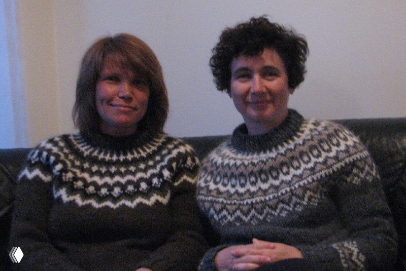 Две женщины в традиционных исландских свитерах сидят в интерьере, показывая плотную текстуру ткани и характерные национальные узоры изделий ручной работы.