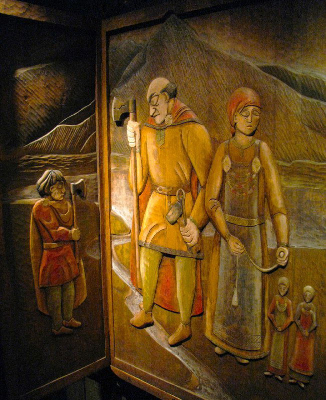 Деревянная резная панель с изображениями викингов и сценами из саги: музейный экспонат с выраженной рельефной работой и тёплой древесной фактурой.