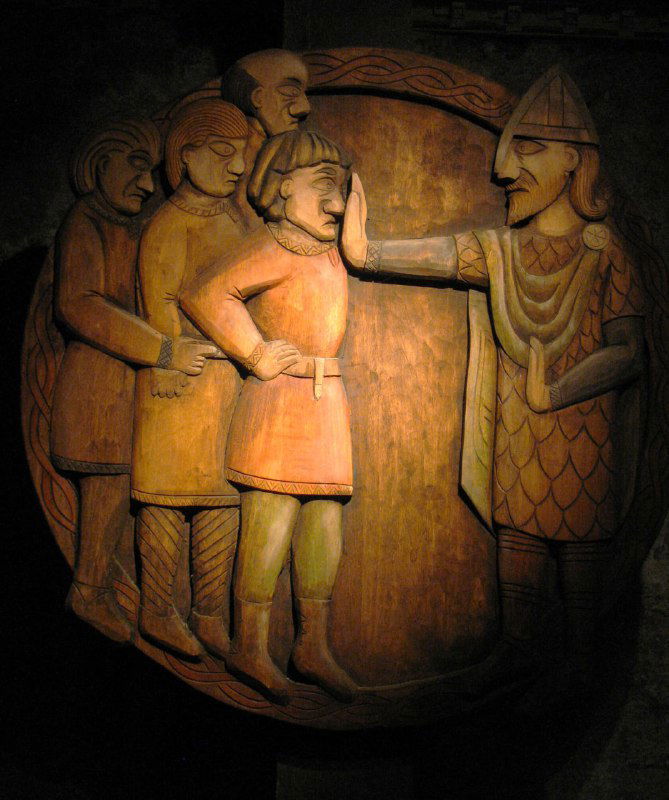 Крупный план резного фрагмента саги об Эгиле: лица персонажей и сцены конфликтов, чёткая детализация на тёмном фоне музейной витрины.