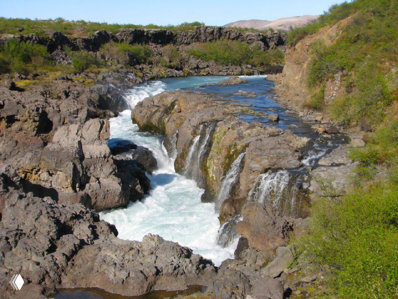 Узкий каскадный водопад в каменном ущелье с бурлящей рекой и порогами, образующими прозрачные водовороты и пенящиеся потоки между скалами.