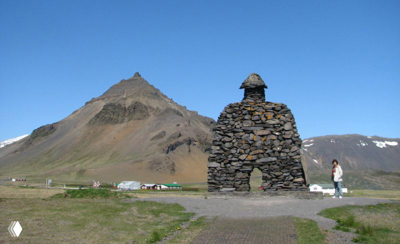 Каменное изваяние-памятник великану Бардару в Арнастапи на фоне холмов и гор, вид у начала тропы вдоль берега.