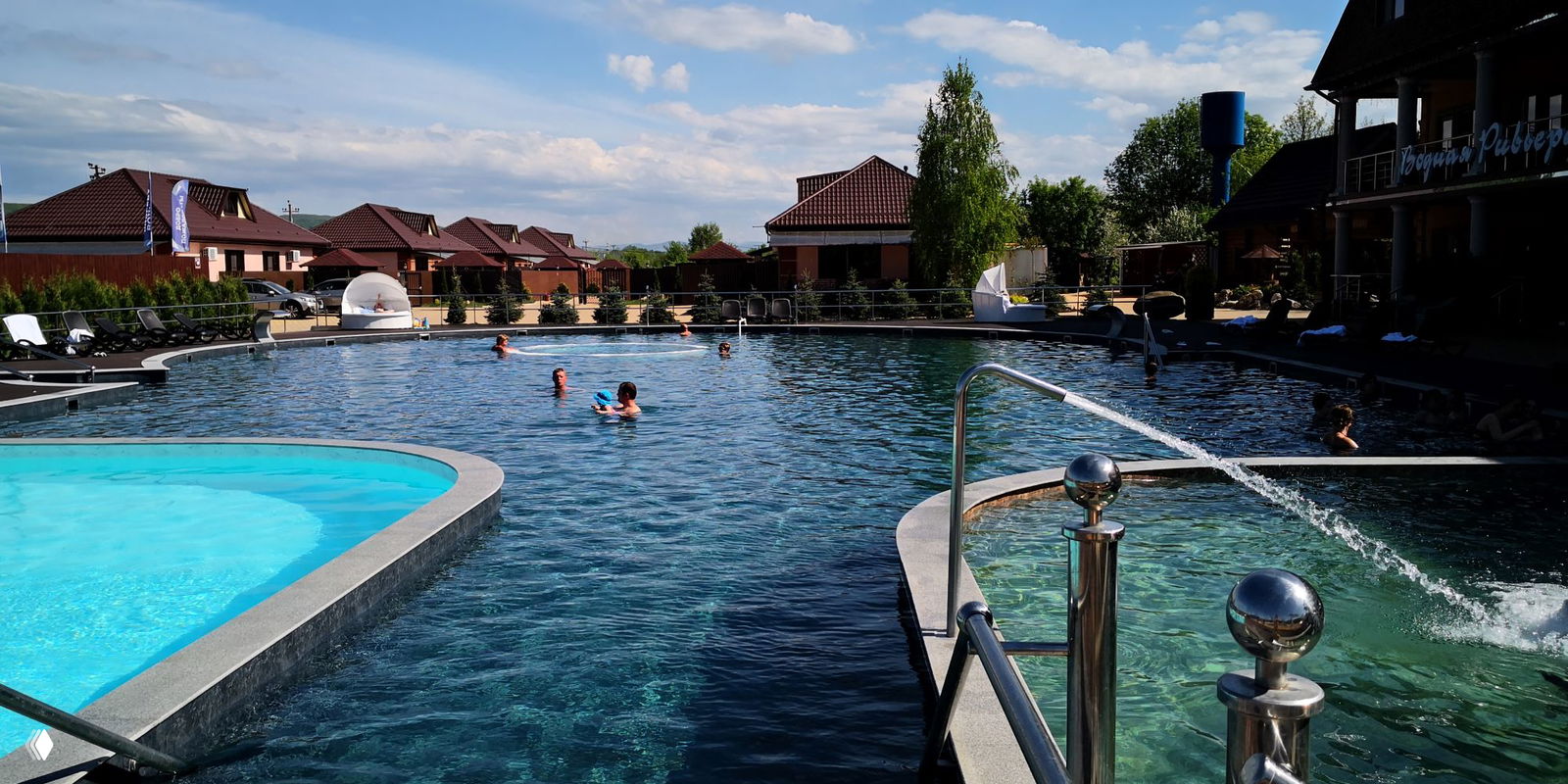 Дневной вид на несколько бассейнов разных температур и причудливые струи воды из металлических фонтанчиков, зона отдыха на фоне.