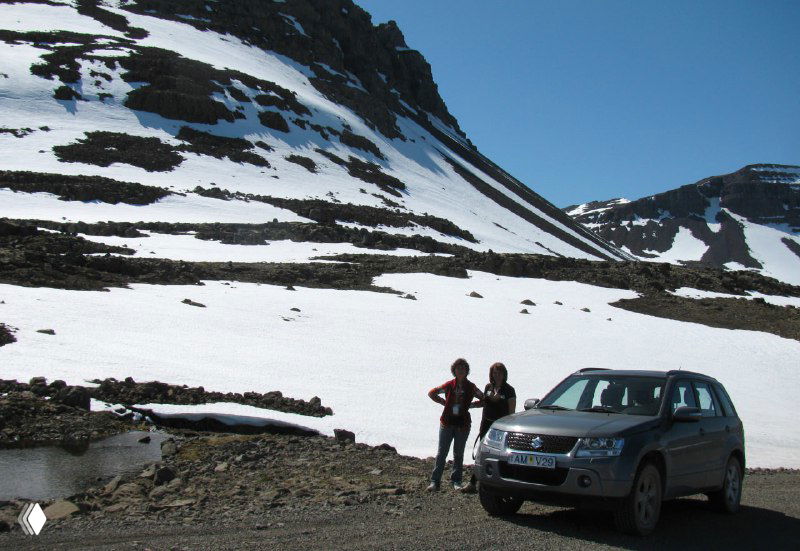 Два путешественника и припаркованный автомобиль на фоне заснеженного горного перевала — начало подъема к водопаду и альпийский пейзаж.