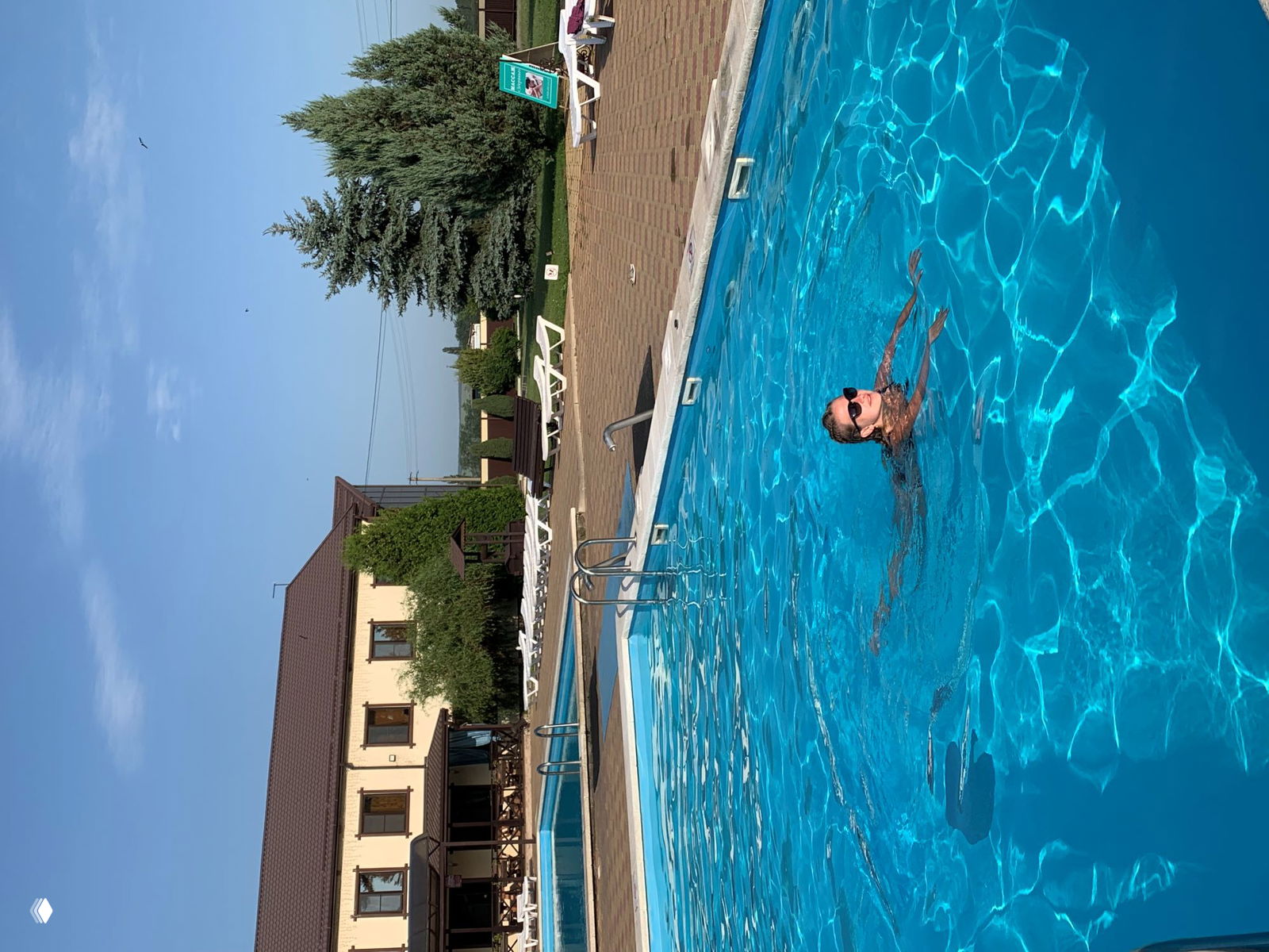 Солнечный кадр открытого бассейна с плавающей девушкой у края, зелёные ели и корпус гостиницы на заднем плане, спокойная сцена отдыха.