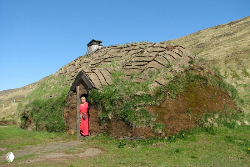 Торфяной дом с густой травой на крыше; у входа стоит человек в яркой одежде — реконструкция традиционного жилища в музее.