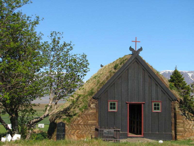 Торфяная деревянная церковь с травяной крышей в исландской деревушке: аккуратный фасад, зелёная кровля и окружающая растительность у подножия гор.