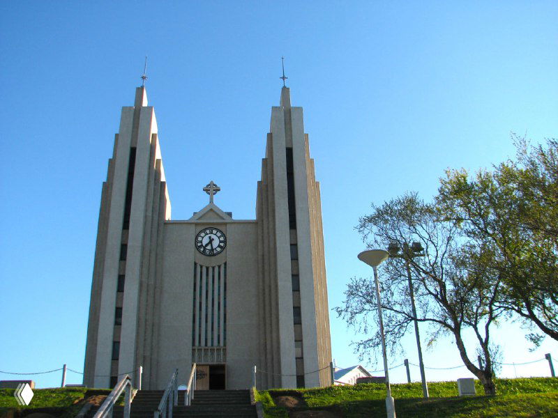 Внушительный собор в городке Акурейри с высокими башнями и лестничным подъёмом: классический фасад, часы и деревья на переднем плане.