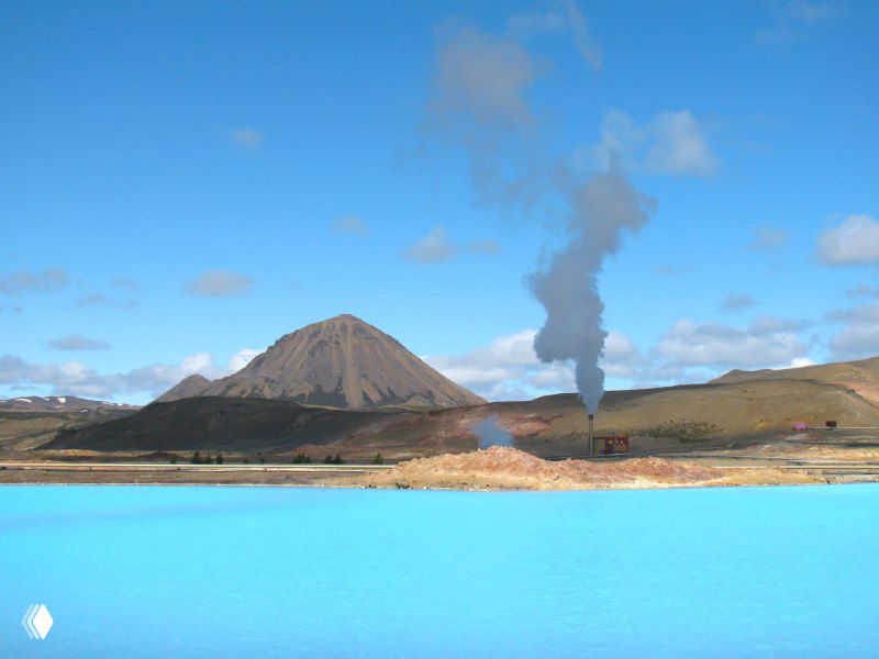 Ярко-голубая термальная лагуна у озера Миватн: насыщенный цвет воды, пар над источниками и вулканический холм на заднем плане в спокойном ландшафте.
