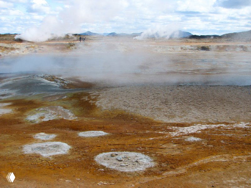 Широкая термальная равнина с паром и разноцветными отложениями: охристые, серые и белые пятна земли, вдалеке холмы и рассеянные облака пара.
