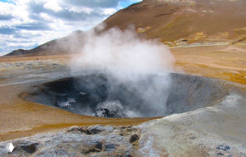 Крупный дымящийся котёл в термальной зоне: темная вода и пар, вокруг обломки застывшей породы и обожжённые окрасы поверхности.