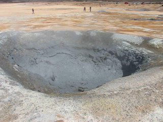Глубокий котёл серой воды на термальной поверхности: ободок вокруг отверстия, в котором видны пузырящиеся потоки и пар, люди далеко для масштаба.