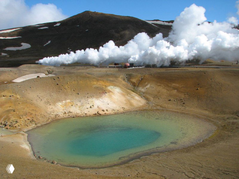 Кратер с бирюзовым озером у подножия холмов Крафлы: яркая вода, желтоватые минералы вокруг и струящийся пар вдалеке на горизонте.