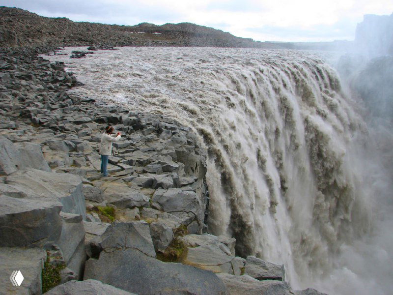 Мощный поток водопада Детифосс с бурной струёй, вид с обзорной площадки и каменные глыбы у кромки падения воды.