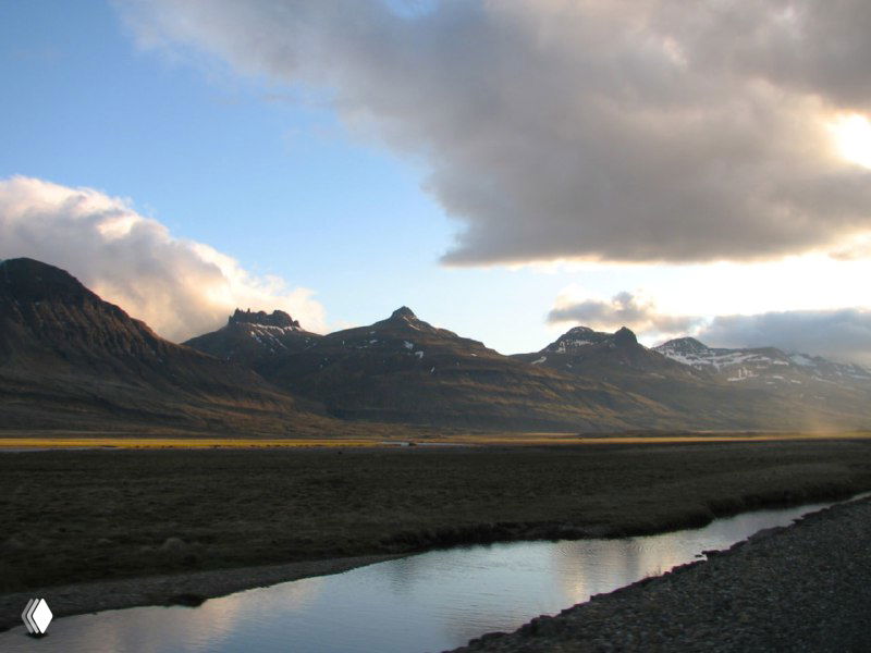 Речка в предгорной долине на фоне силуэтов гор и низкого облачного слоя — спокойный пейзаж вдоль дороги.
