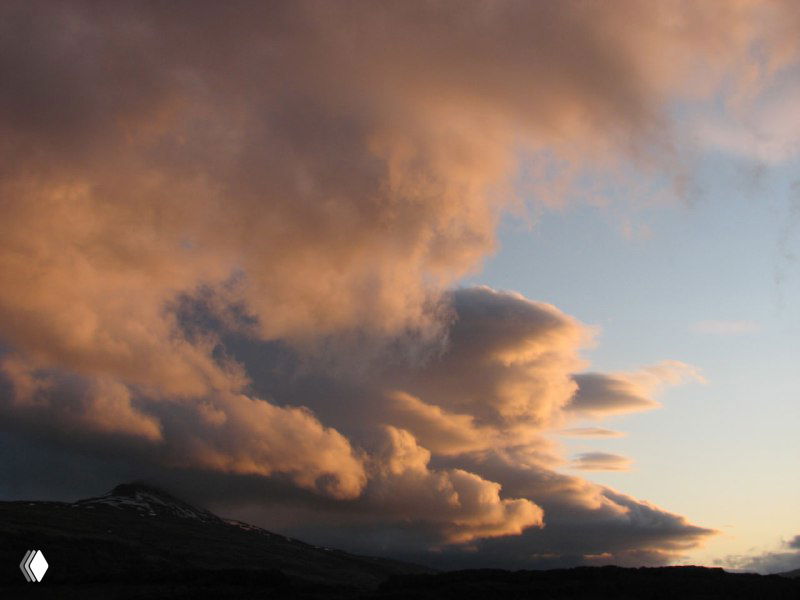 Облака на закате образуют драматичные формы над горным горизонтом, тёплые оттенки неба и контраст темных скал.