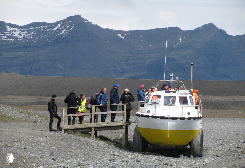 Берег лагуны с пришвартованной лодкой-амфибией и группой туристов, готовых отправиться на экскурсию среди айсбергов.