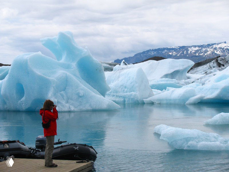 Фотограф на причале снимает огромные льдины; рядом лодочка и небольшие айсберги у кромки воды лагуны.