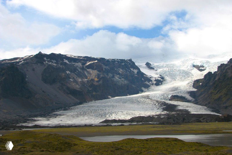 Язык ледника, спускающийся между тёмными скалами; видно контраст льда и вулканической породы в долине.