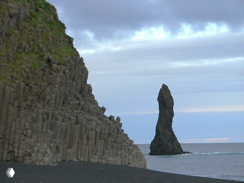Базальтовые колонны на берегу и одиночная высокая скала-валун у моря возле Рейнисдрангар — четкая геологическая структура на фоне неба.