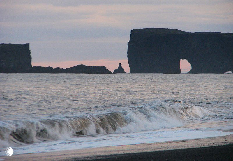 Каменная морская арка мысa Дирхолей и прилегающие скалы, снятые с берега — вид на арочный проход и волны у основания образований.
