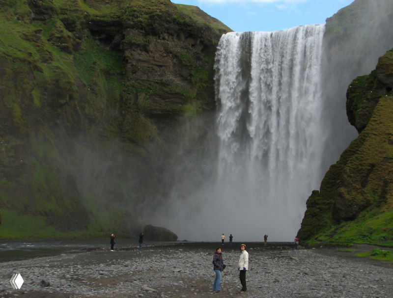 Водопад Скогафосс с 60-метровой струёй воды, падающей с зелёного утёса; внизу у подножия видны люди для масштаба и брызги.