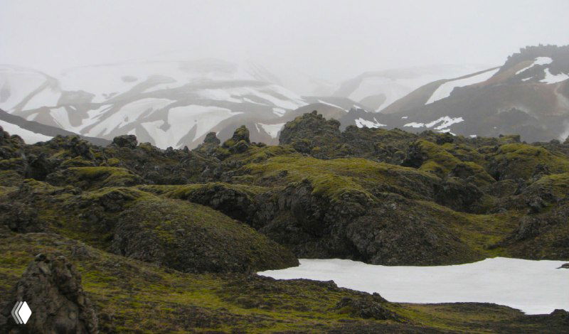 Моховые холмы и кратероподобные образования на фоне туманных заснеженных вершин — суровый и зелёный исландский ландшафт.