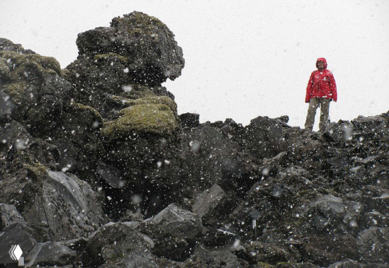 Человек в красной куртке стоит на чёрных лавовых валунах, вокруг падает снег — контрастная сцена похода в суровой погоде.