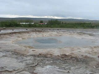 Спокойная серо-коричневая лужа гейзера на площадке: место, откуда периодически вырывается фонтан воды, окружающий сельский пейзаж.