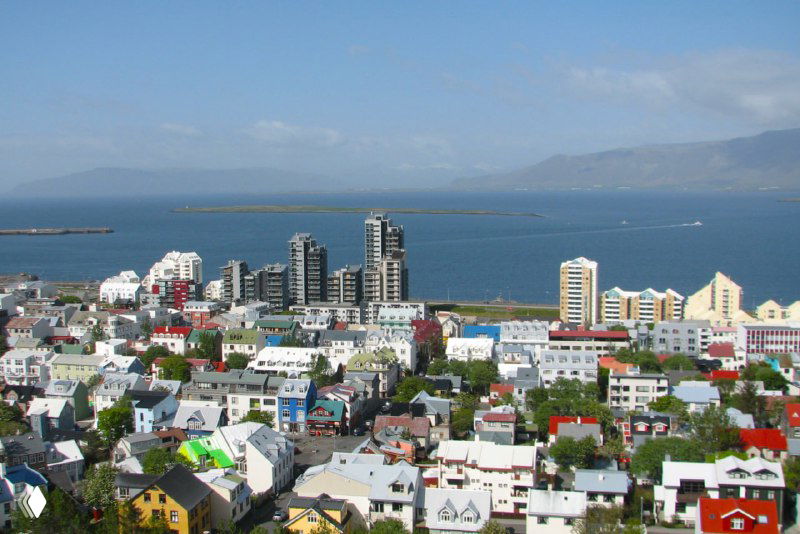 Панорамный вид на жилые кварталы Рейкьявика, разноцветные крыши и побережье вдалеке — городской ландшафт у моря.