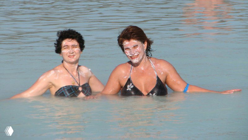 Две женщины купаются в голубой воде термального комплекса, одна из них с белым косметическим налётом на лице — сцена отдыха.