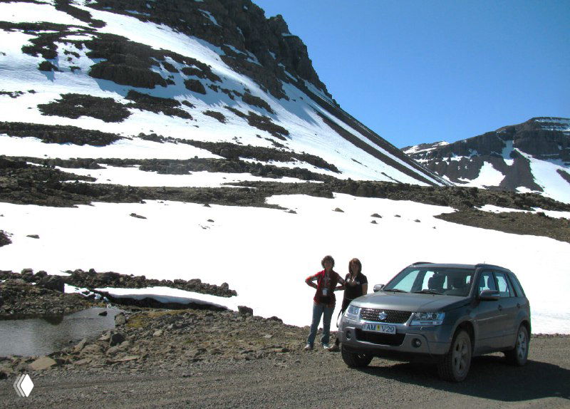 Внедорожник на горной грунтовке у снежных склонов; рядом два человека у машины — показывает сложность дорог и масштабы ландшафта в горах.