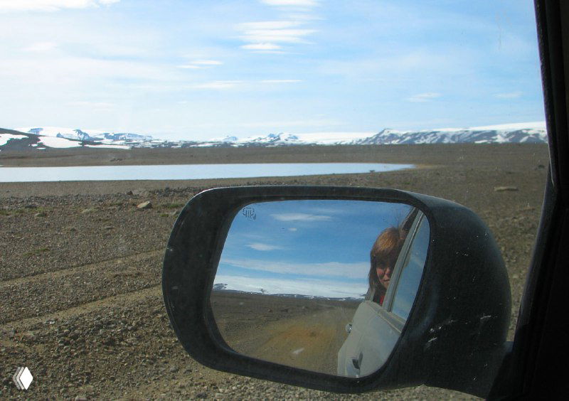 Вид через боковое зеркало автомобиля на пустынную гравийную дорогу, озеро и дальние горы — отражение атмосферы исландской поездки и пути.