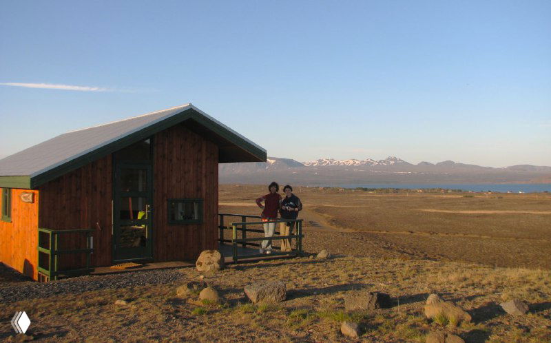 Небольшой деревянный гостевой домик на плато у озера с горным горизонтом, пример бюджетного размещения и уединённого пейзажа региона.