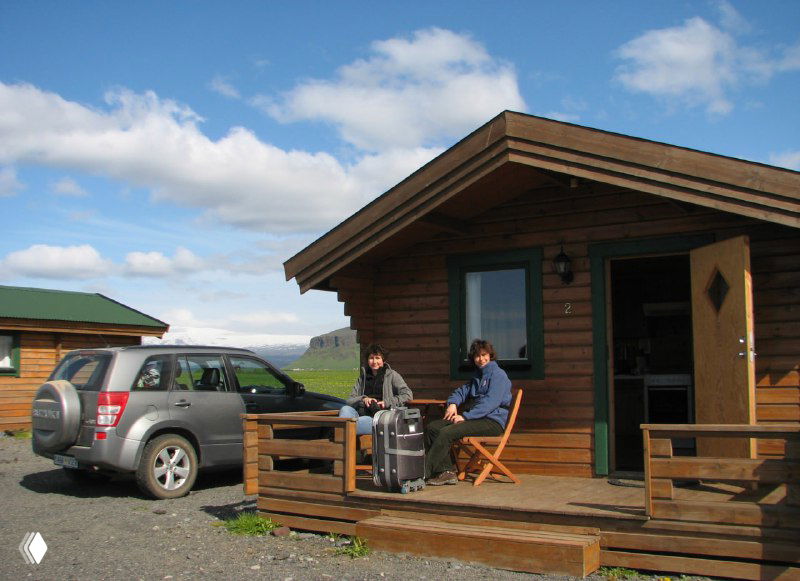 Кемпинговый домик с открытой верандой, рядом чемоданы и автомобиль — сцена размещения и отдыха во время автопохода по Исландии в начале сезона.