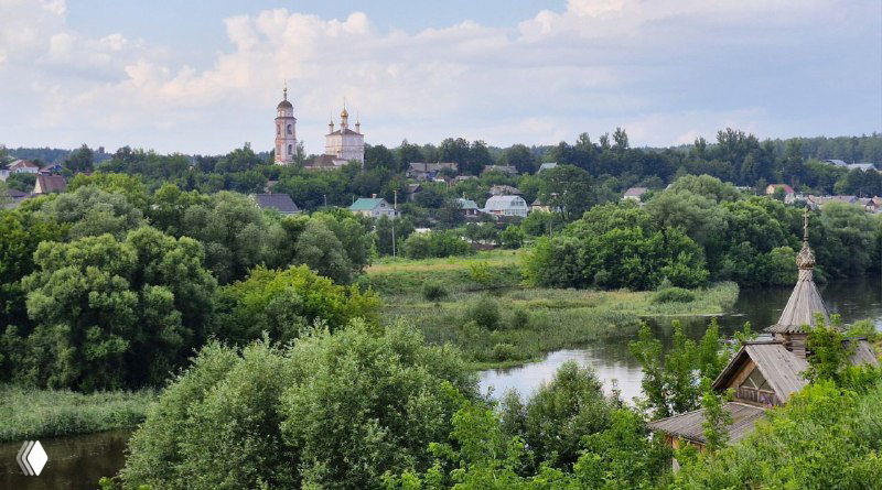 Вид через зелёные луга на высокую церковь в Боровске и деревянные дома у берега, характерный сельский пейзаж района.