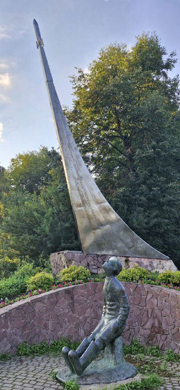 Памятник Циолковскому: высокий стреловидный обелиск и бронзовая фигура учёного в валенках на постаменте среди зелени.