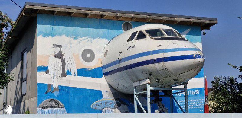 Нос настоящего самолёта Як-40, встроенный в фасад музейного здания, рядом крупная настенная роспись с авиационной тематикой.