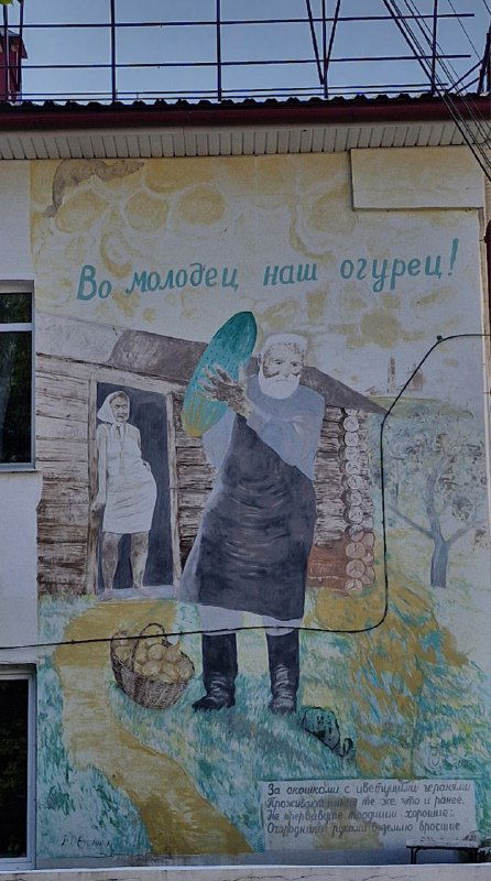 Роспись с надписью «Во молодец наш огурец!» и изображением женщины с корзиной и вёдрами на фоне фасада в центре города