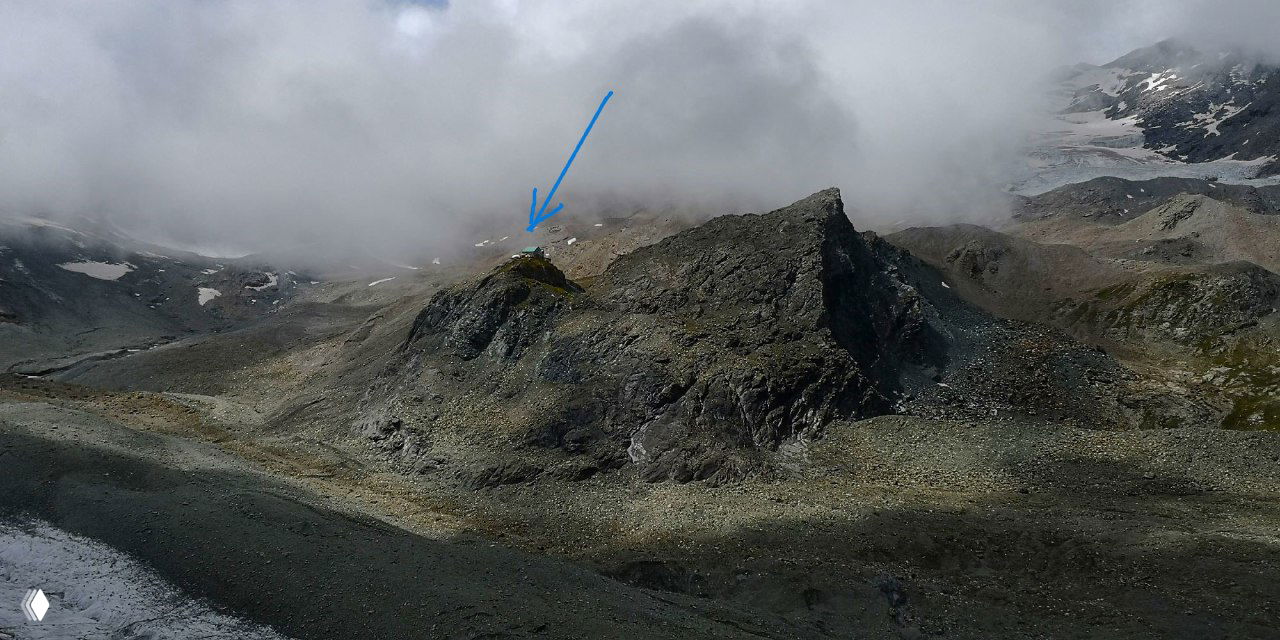 Высокогорный каменистый ландшафт с гребнем и небольшой удалённой хижиной, отмеченной стрелкой, в облачную погоду — пример сложного маршрута между приютами.