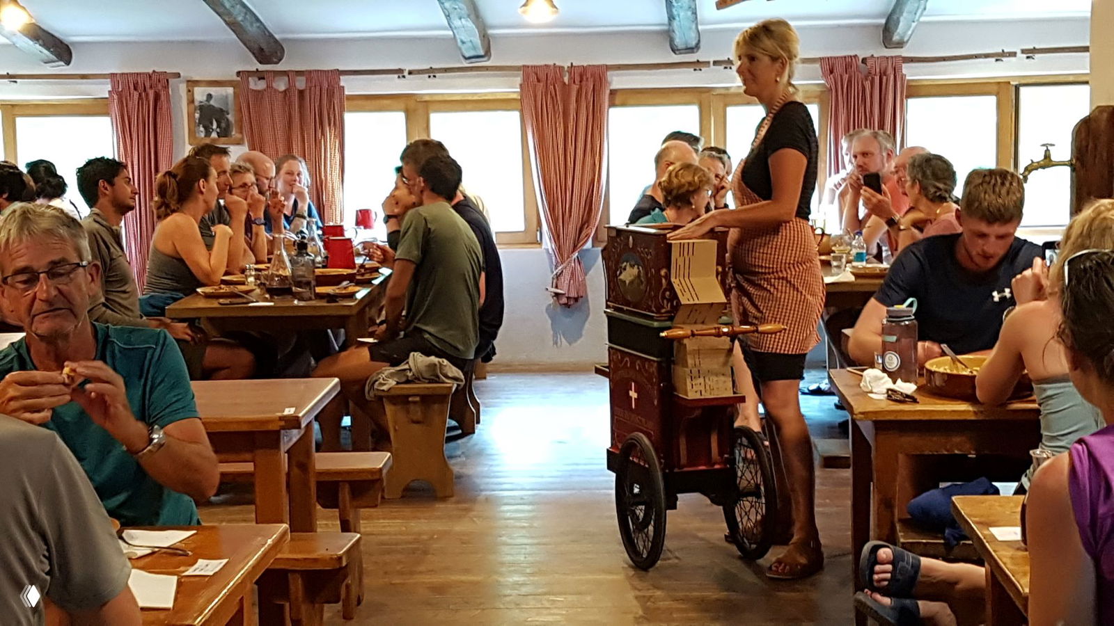 Столовая горного приюта во время общего ужина: гости за длинными столами, атмосфера общения, хозяйка играет на шарманке — вечерняя сцена из жизни приюта.