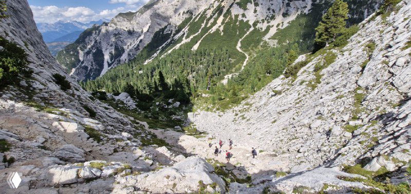 Крутой каменистый спуск по тропе Alta Via, группа туристов спускается по щебенистому руслу между скал.