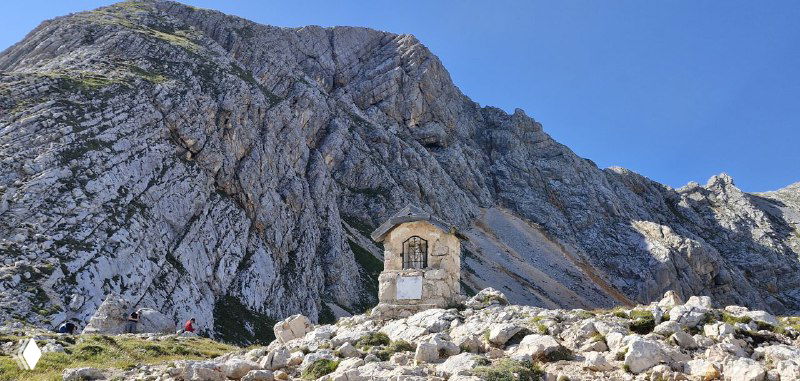 Маленькая каменная часовня или приют на фоне высоких скал Доломитов, каменная постройка среди камней и травы.