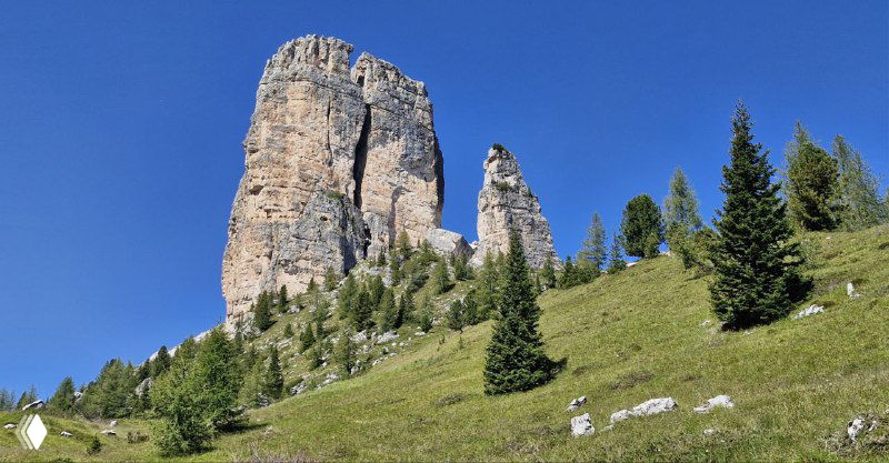 Одна высокая скала Cinque Torri на зелёном склоне; хвойные деревья у подножия и яркое голубое небо над вершиной.